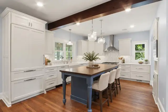 Remodeled kitchen with white inset cabinets, navy island with wood top, dark wood ceiling beam and glass pendant lights