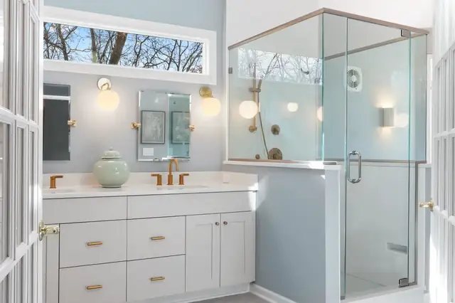 Bathroom with white shaker double vanity, round brass sconces, clerestory window and glass corner shower