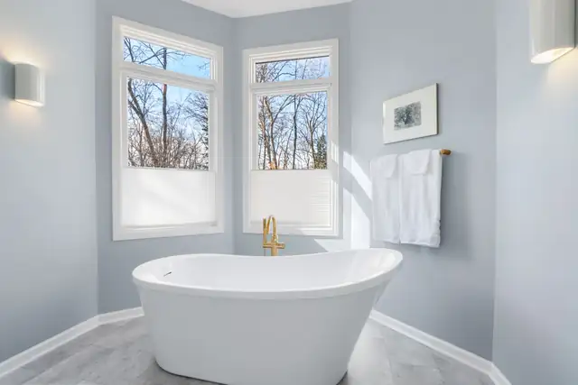 Bright bathroom with freestanding soaking tub, brass floor-mounted filler, corner windows and gray tile floor