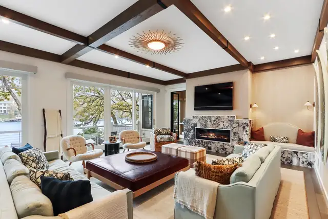 Lake home living room with dark wood coffered beams, marble fireplace surround, sunburst light and water view